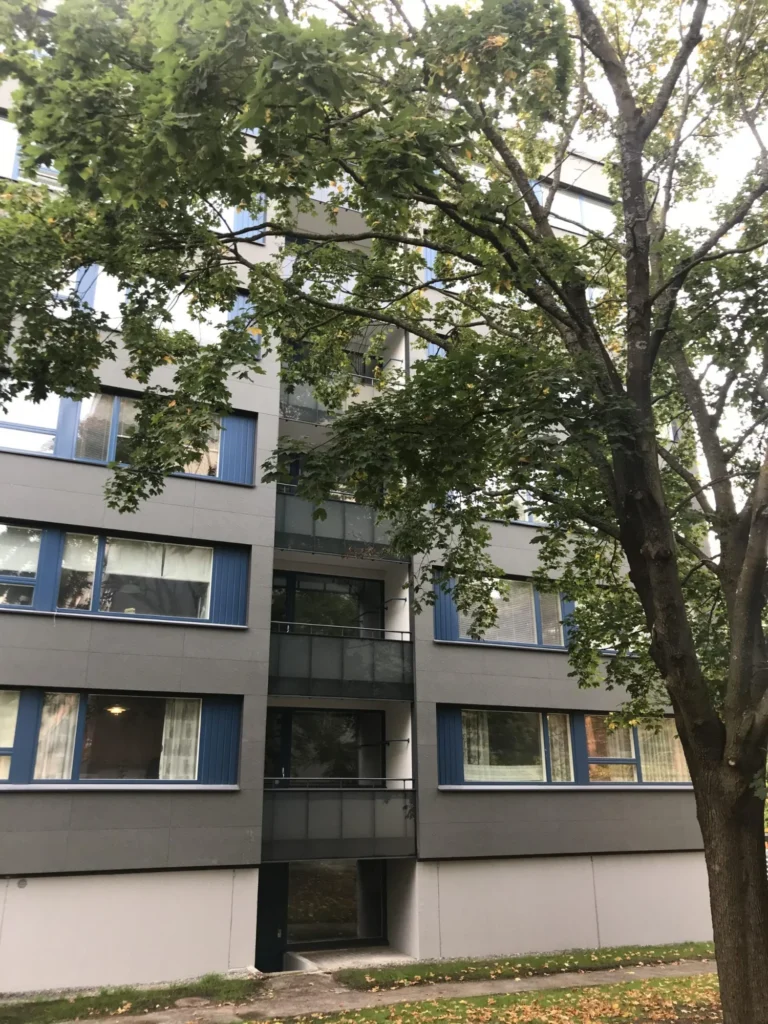 As Oy Nunnanhaka parvekejulkisivu, harmaat julkisivulevyt, siniset ikkunat ja puu rakennuksen edessä