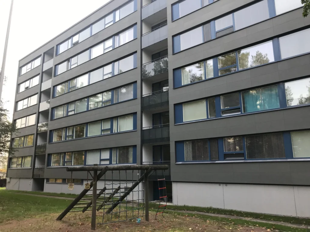 As Oy Nunnanhaka pihan puolelta, harmaat julkisivulevyt, siniset ikkunapuitteet ja leikkipaikka kerrostalon edessä