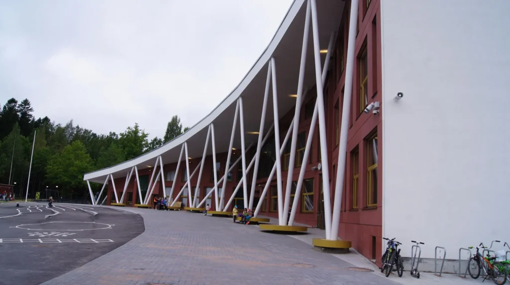 Auroran koulun kaareva valkoinen katos ja vinot teräspilarit punaisen concrete skin -julkisivun edessä, Espoo