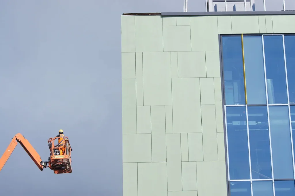 Asentaja henkilönostimessa kiinnittämässä vihreitä concrete skin -levyjä Herttoniemen metrovalvomon julkisivuun, Helsinki