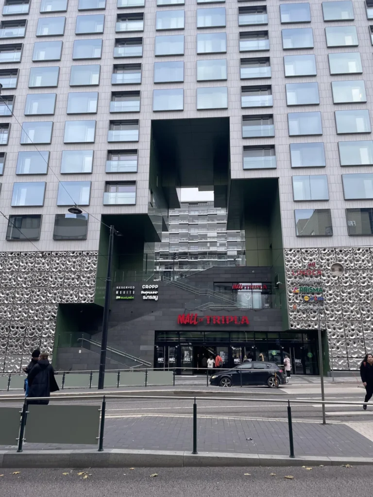 Laaja näkymä Mall of Triplan katujulkisivusta, pääsisäänkäynnistä, viherreunaisesta rakennusaukosta ja vaaleista kuviopaneeleista Helsingissä