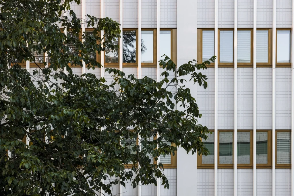 Ratamestarinkatu 9 julkisivun yksityiskohta, valkoinen ULMA levypinta, pystyrimat ja kullanväriset ikkunat puuston takana